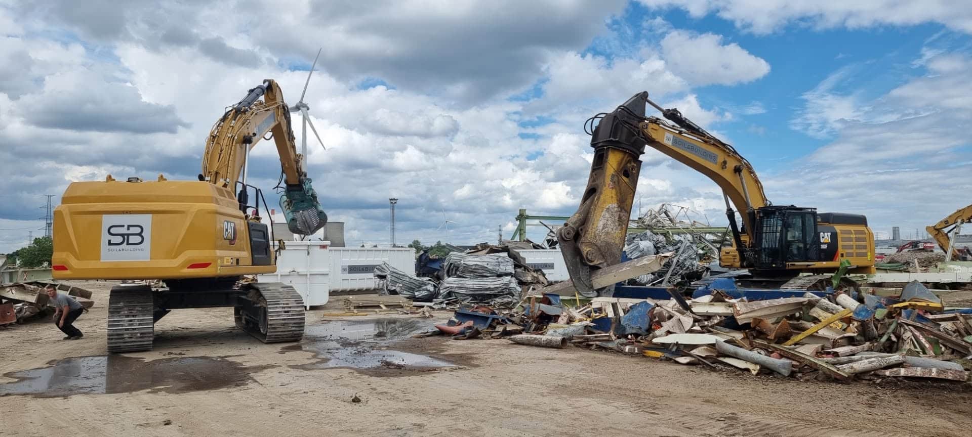 Twee gele CAT rupskranen van Soil & Building in actie op een sloopwerf, omringd door puin en schroot, met windturbines op de achtergrond