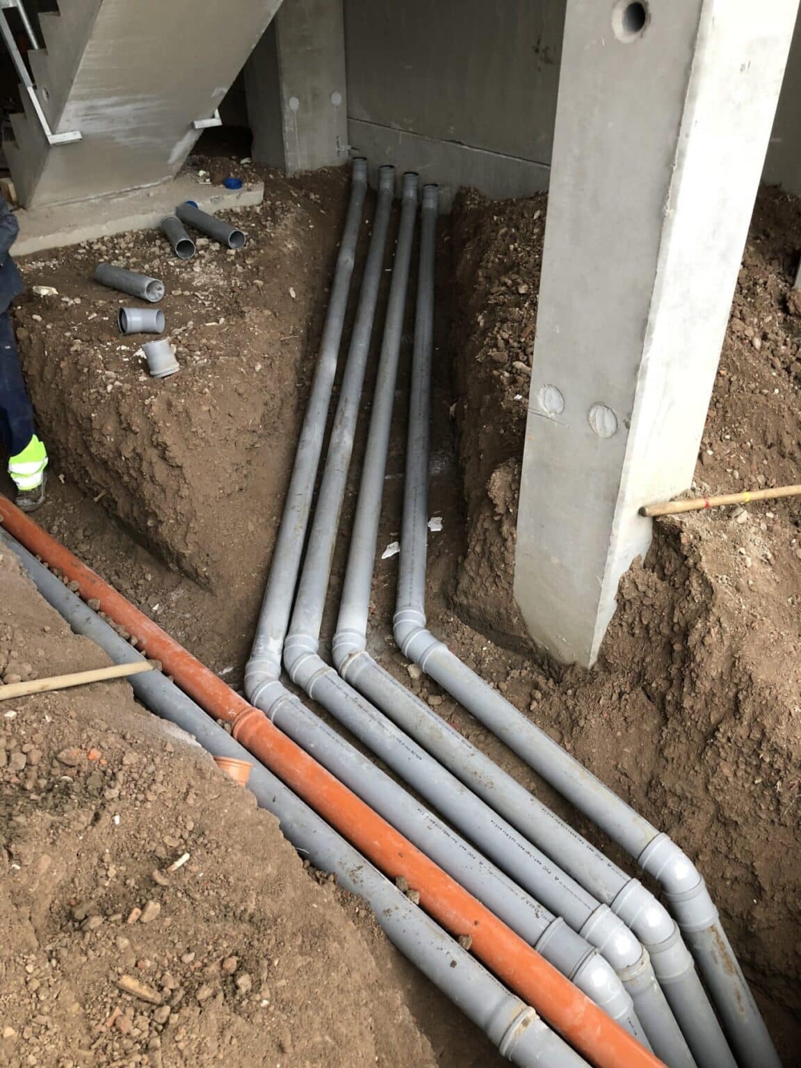 Grijs en oranje PVC-afvoerbuizen voor regen- en vuilwater in een bouwput naast een betonnen kolom op MG Park De Hulst