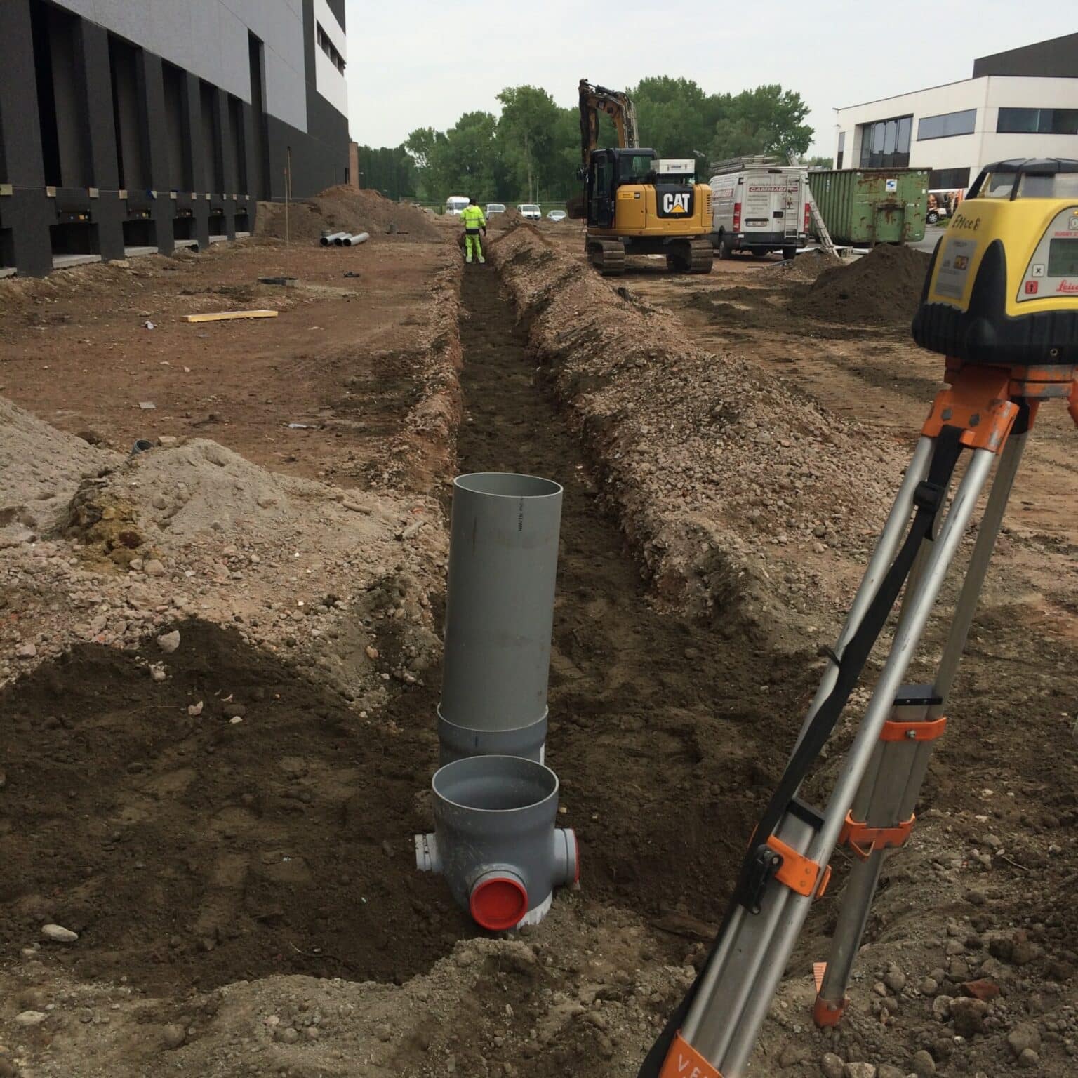 Rioleringswerken in sleuf op de DHL-werf in Bornem met CAT-graafmachine, Leica meetapparatuur en PVC-buizen