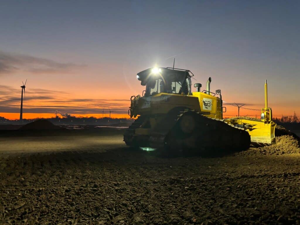 Caterpillar bulldozer van Soil & Building egaliseert een terrein bij zonsondergang, met windturbines op de achtergrond