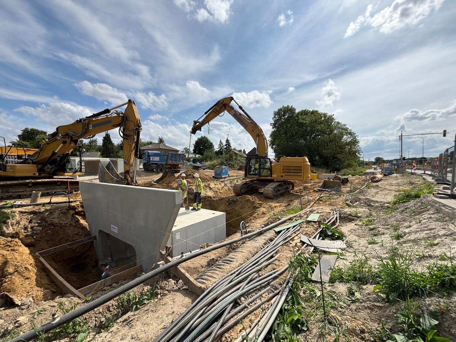 Twee rupsgraafmachines bij grondwerken op een infrastructuurwerf, met betonnen constructie-elementen en werklieden