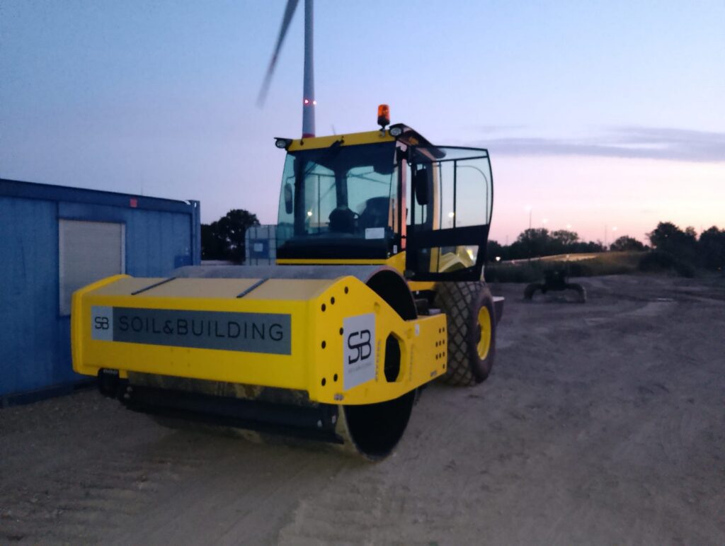 Gele wals van Soil & Building op een bouwwerf bij schemering, met een windturbine op de achtergrond