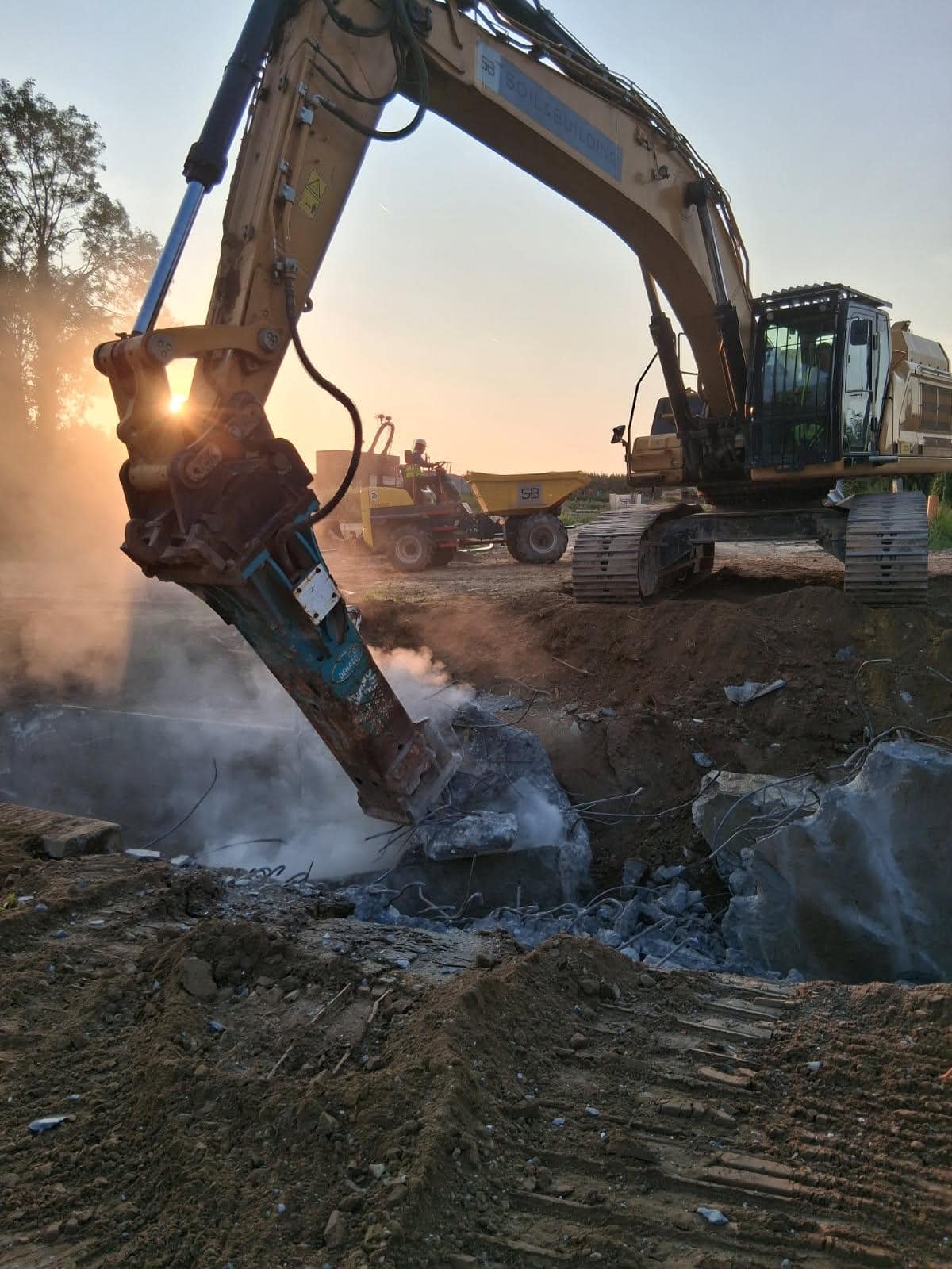 Rupsgraafmachine van Soil & Building met hydraulische hamer die betonnen constructie breekt op een werf, omgeven door stof