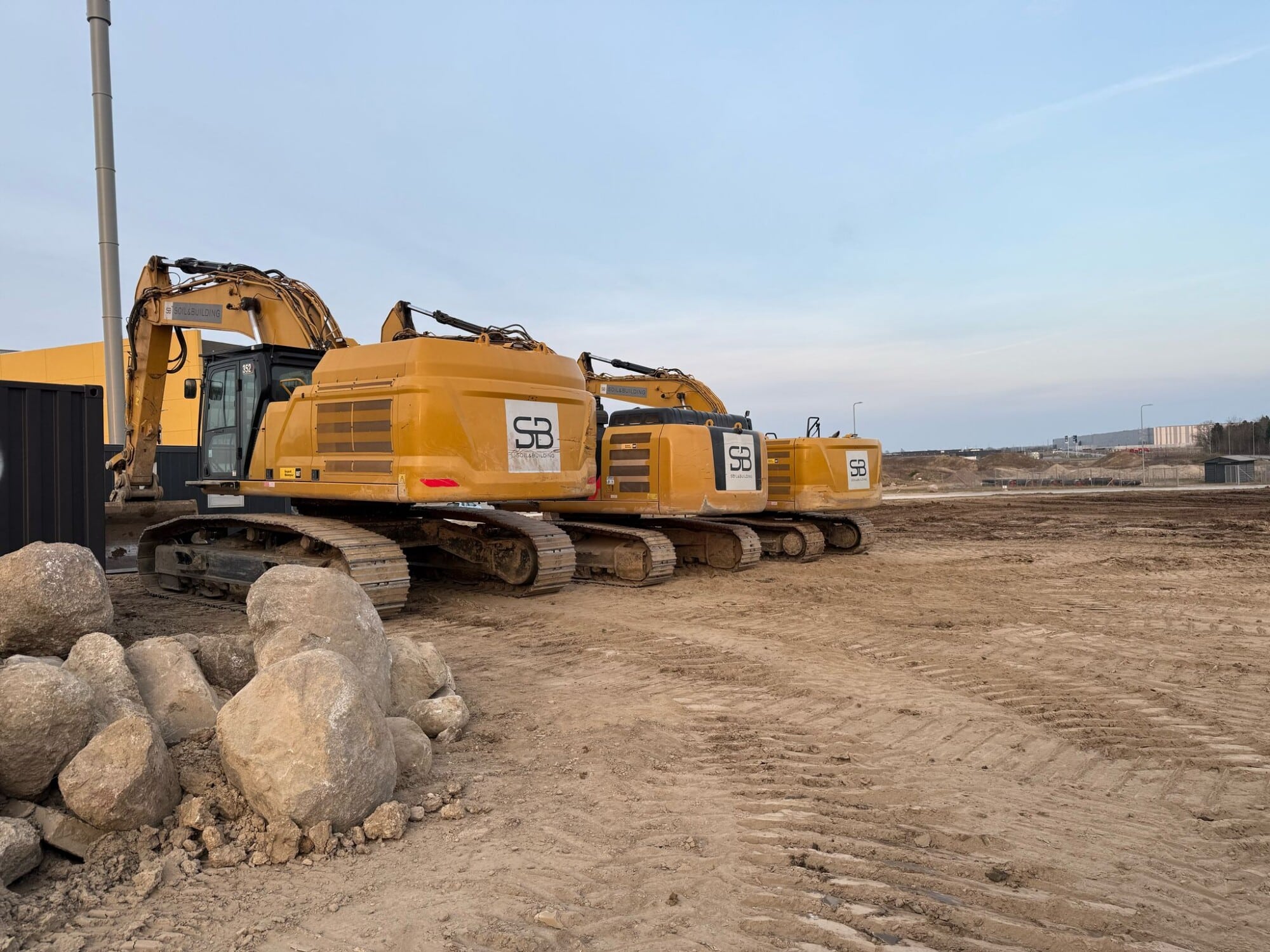 Drie gele rupsgraafmachines van Soil & Building op een bouwterrein met grote stenen op de voorgrond