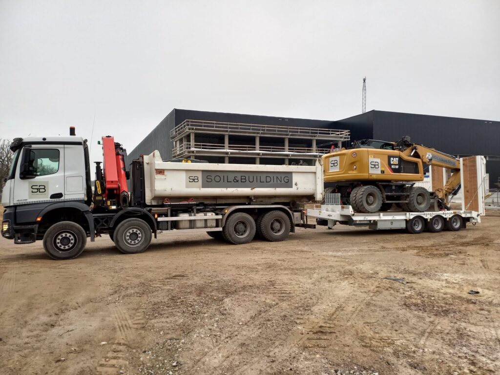 Vrachtwagen met autolaadkraan van Soil & Building vervoert een mobiele graafmachine op een aanhangwagen met verlaagd dek naar een bouwwerf